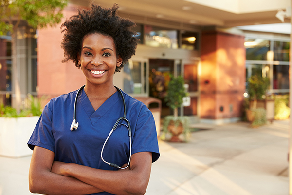 Confident female nurse in blue scrubs standing outside a healthcare facility with arms crossed and smiling