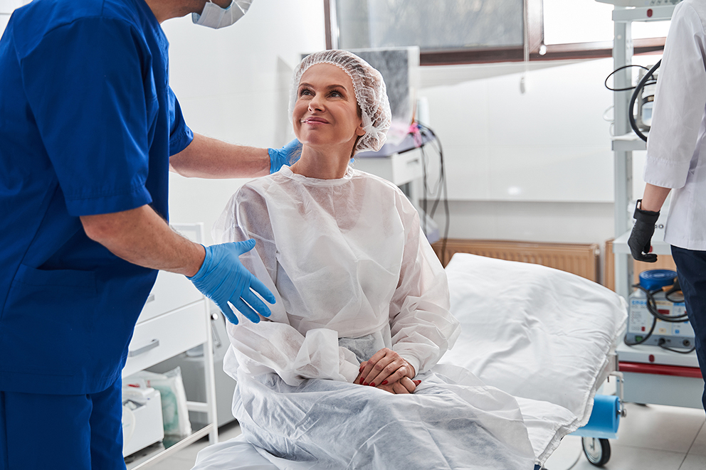 Patient in a surgical gown sitting on a hospital bed, smiling and talking with a medical professional before a procedure